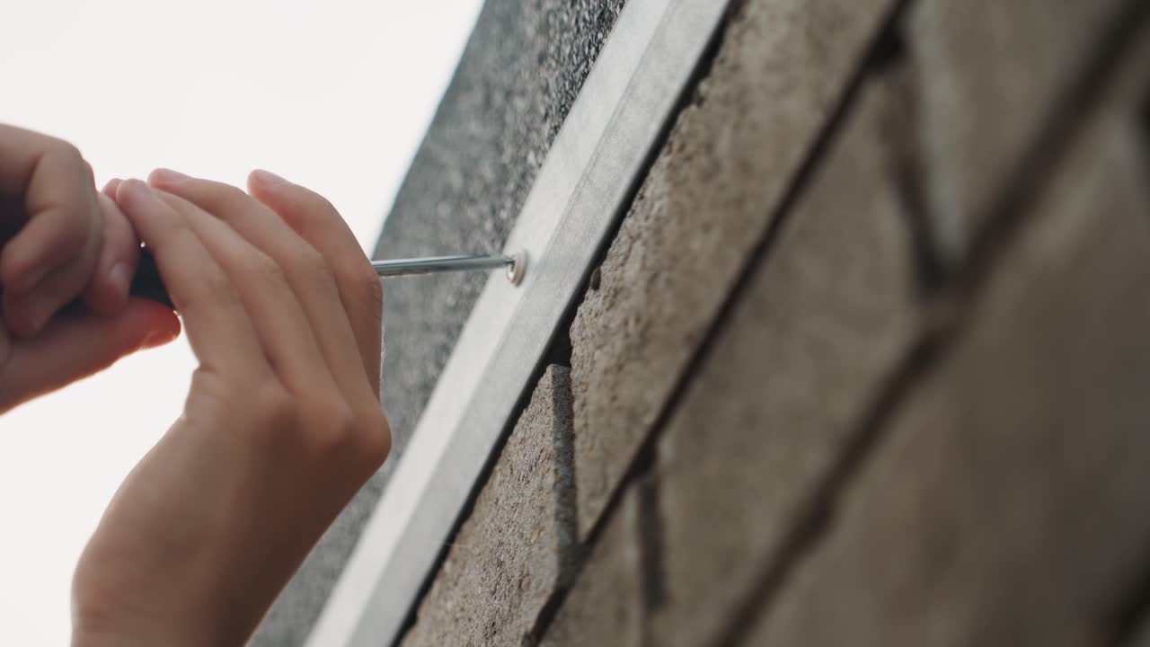 Dichte opname van handen die metalen daktrim vastschroeven, bakstenen muur als achtergrond onder bewolkte hemel, geconcentreerde huiseigenaar gebruikt kleine schroevendraaier, nauwkeurige uitlijning van de strook, tastbare textuur, kalme weloverwogen reparatie