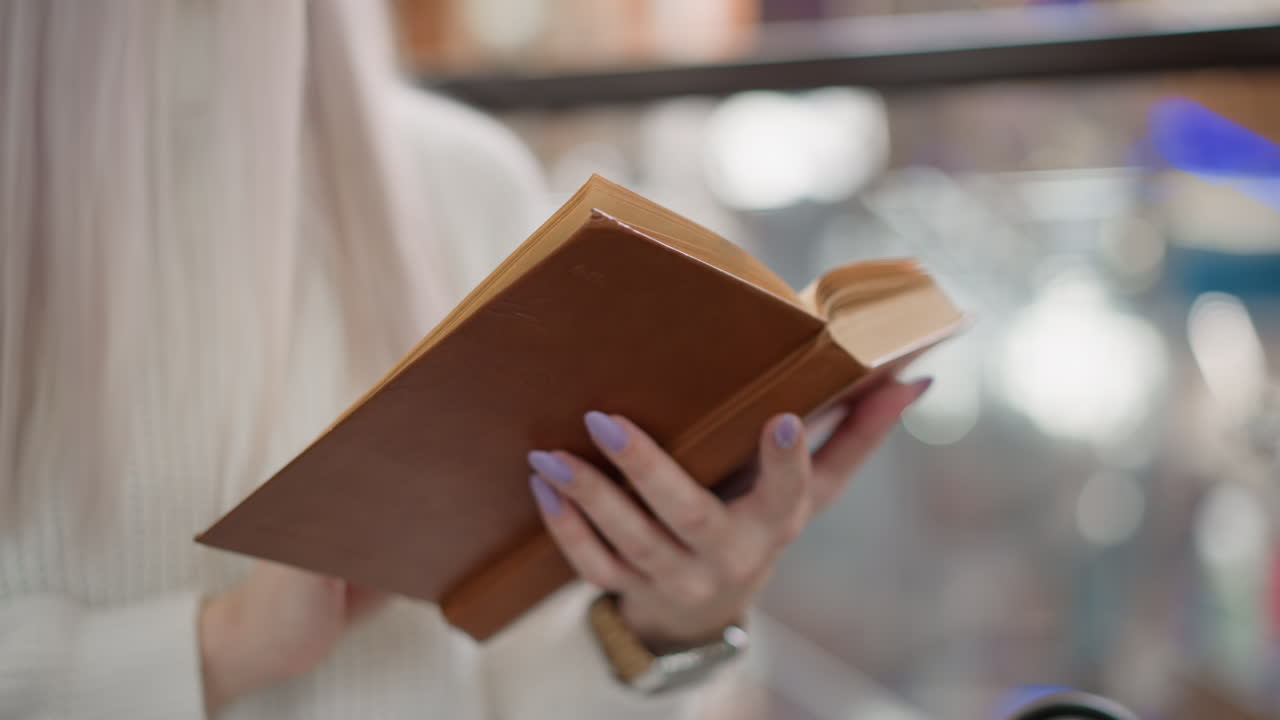 lower view of youthful woman hand flipping pages of brown bound novel on mall bench with pastel nails blur bokeh background cozy reading moment wearing white sweater wristwatch black bag beside