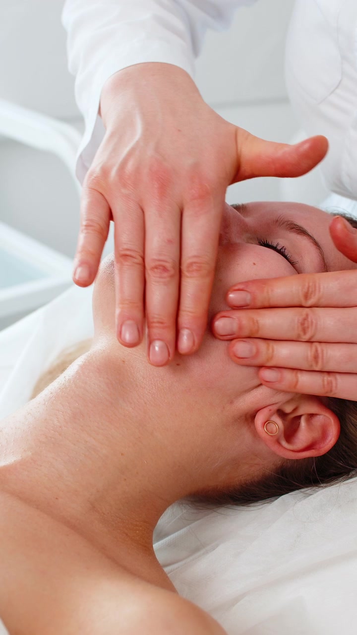 Ung kvinde får kæbelinje-ansigtsmassage af kosmetolog i et moderne, afslappende spa- og kosmetologiklinikrum