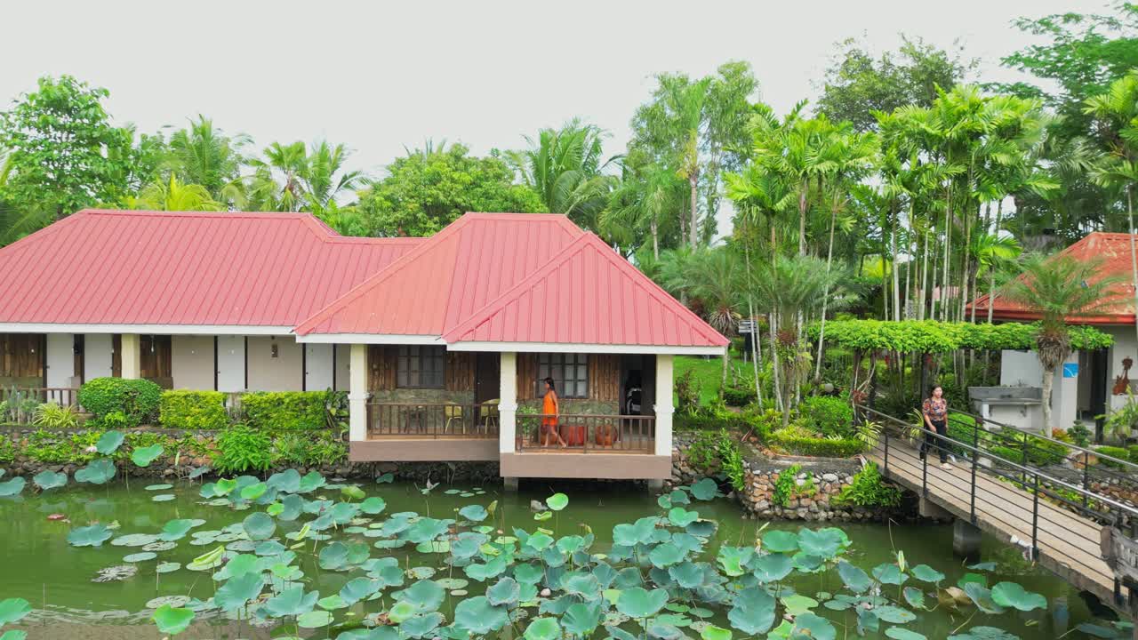 Cảnh quay rộng một người phụ nữ mặc váy cam đi dạo trên ban công phòng nhà gỗ nổi tại Khách sạn Palaisdaan, tọa lạc trên một cái ao đầy lá sen ở Bagabag, Nueva Vizcaya, Philippines.