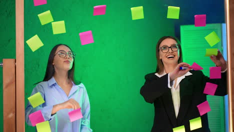cheerful ladies in the office take away stickers from the glass board. business, startup, planning, management and people concept.