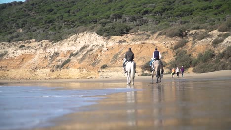 disparo manual en ángulo bajo en la playa con olas tranquilas en el mar azul con vista de dos jinetes con sus caballos cabalgando por la playa
