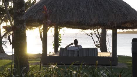 Single-woman-enjoys-tropical-sunset-in-resort-cabana-at-beach-in-Fiji,-dolly-in