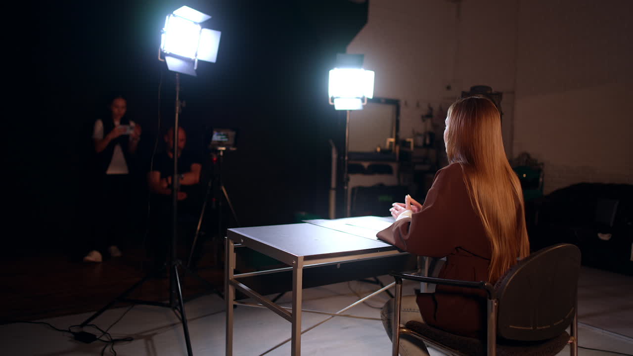 Rear view of a brunette long-haired woman recording a video in studio. Male cameraman and female assistant work backstage.