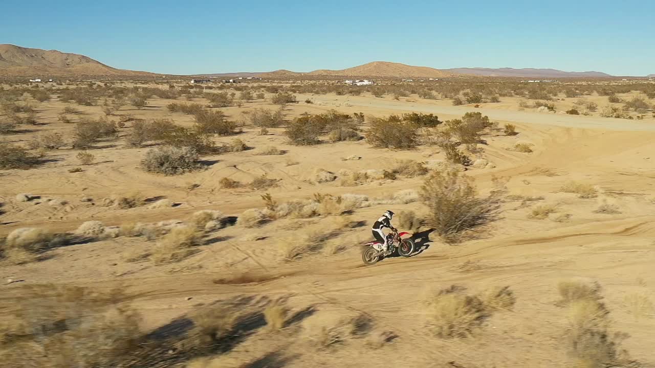 motociclista recorriendo un camino de tierra lleno de baches en el desierto de mojave y saltando un camino en cámara lenta - vista aérea