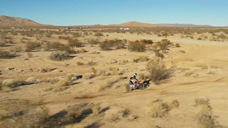 motorcyclist riding along a bumpy dirt trail in the mojave desert and jumping a road in slow motion - aerial view