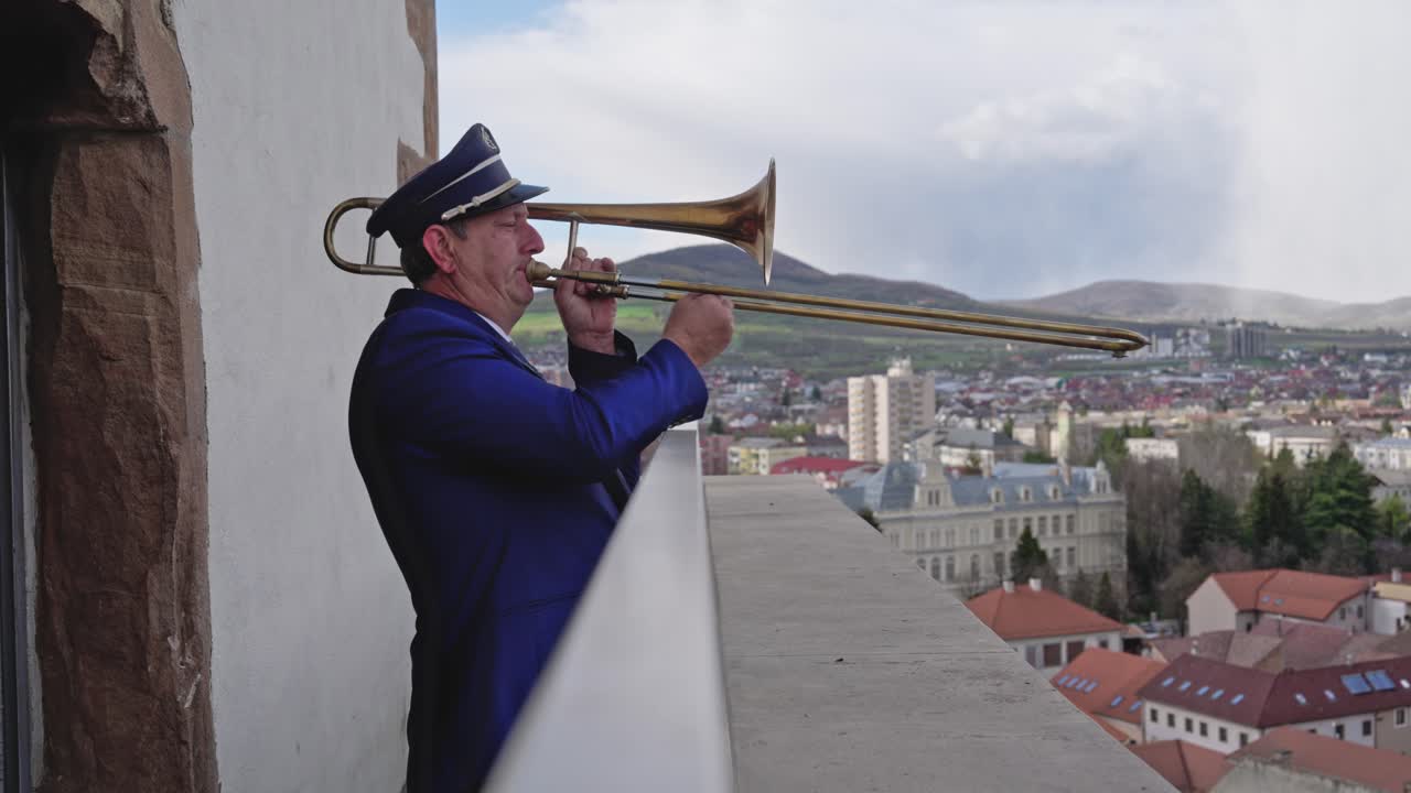 A static shot shows a middle aged man in a navy uniform and hat passionately playing a brass trombone. The background is a bright, scenic view of a European city on a cloudy day