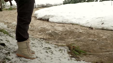 Hiker-standing-near-a-mountain-river-in-a-snow-covered-landscape