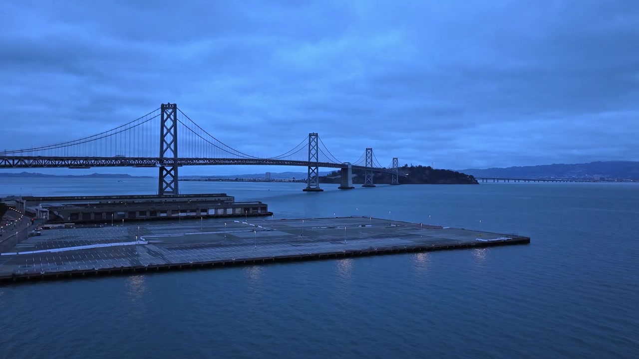 Ripresa ascendente con drone del Ponte della Baia di San Francisco al crepuscolo, con illuminazione dell'ora blu, acqua calma ed edifici illuminati sul lungomare sotto un cielo nuvoloso. Perfetto per progetti di viaggio o paesaggi urbani