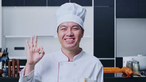 un primer plano de un cocinero asiático sonriendo y mostrando un gesto de acuerdo a la cámara mientras está de pie en la cocina de casa