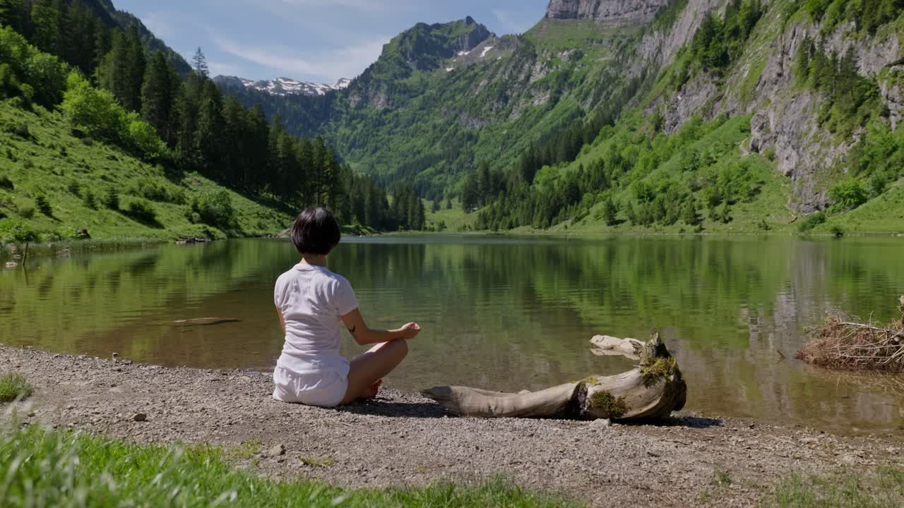 Woman sits by Talalpsee lake in Filzbach, surrounded by mountains and lush greenery, meditating