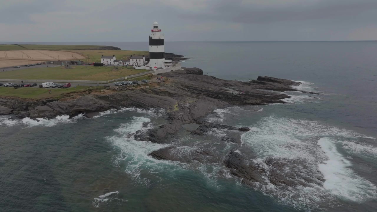 Et luftperspektiv af det historiske sort-hvide Hook Fyr, der står på en barsk klippefyldt halvø med brusende havbølger i County Wexford, Irland.