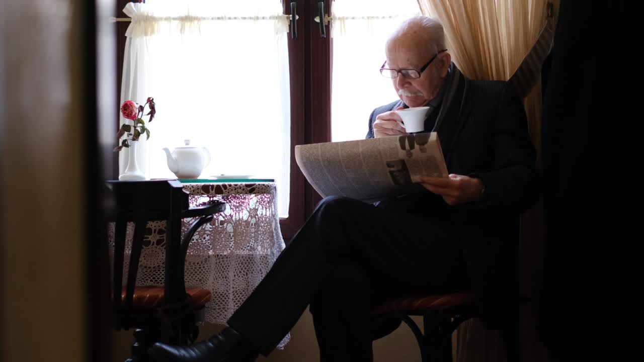 Gray-haired grandfather is sitting in a cafe drinking tea, he is reading a newspaper.