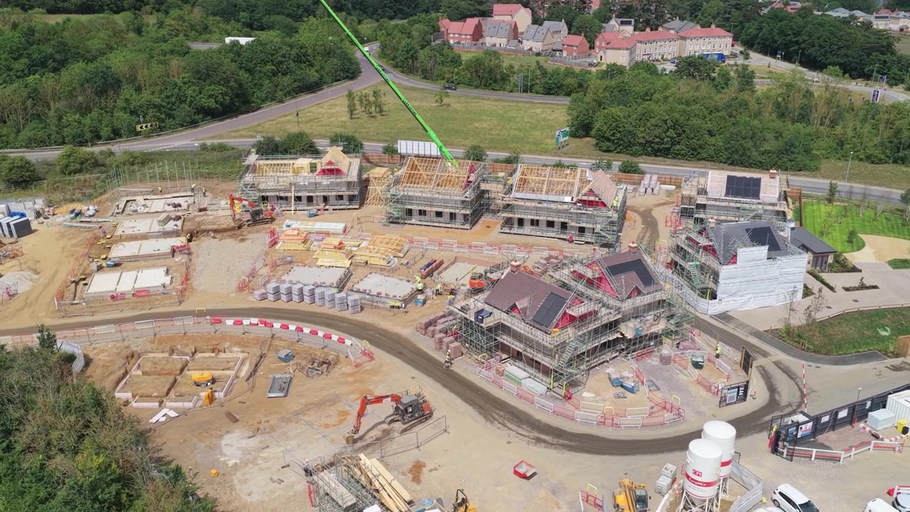 Case residenziali in costruzione con impalcature e macchinari visibili sul cantiere durante un'ora diurna limpida in Inghilterra, con campi verdi che circondano l'area.