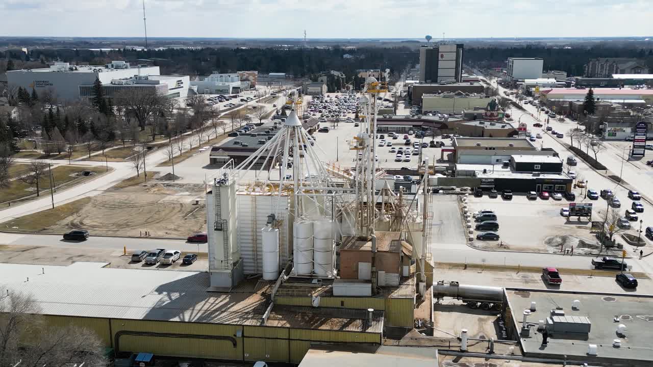 Ripresa aerea rotante con drone di un grande impianto chimico industriale e edificio di fabbrica situato in una piccola città, circondato da attività commerciali locali, strade e auto di passaggio durante il giorno