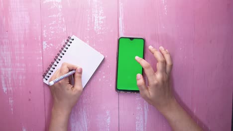 vista superior de la mano de una mujer usando un teléfono inteligente y escribiendo en un bloc de notas