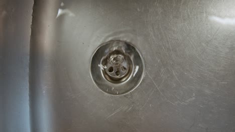 close up of metal kitchen sink washbasin full of water draining through hole with swirling motion in slowmo