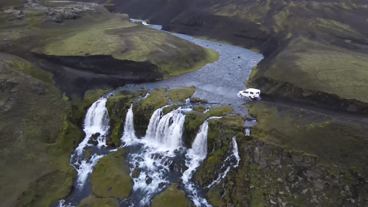 Vista aerea da drone di una cascata impetuosa accanto a una strada di montagna tortuosa con un veicolo bianco che passa. Il paesaggio montuoso remoto mostra fiumi, scogliere e natura aspra in Islanda