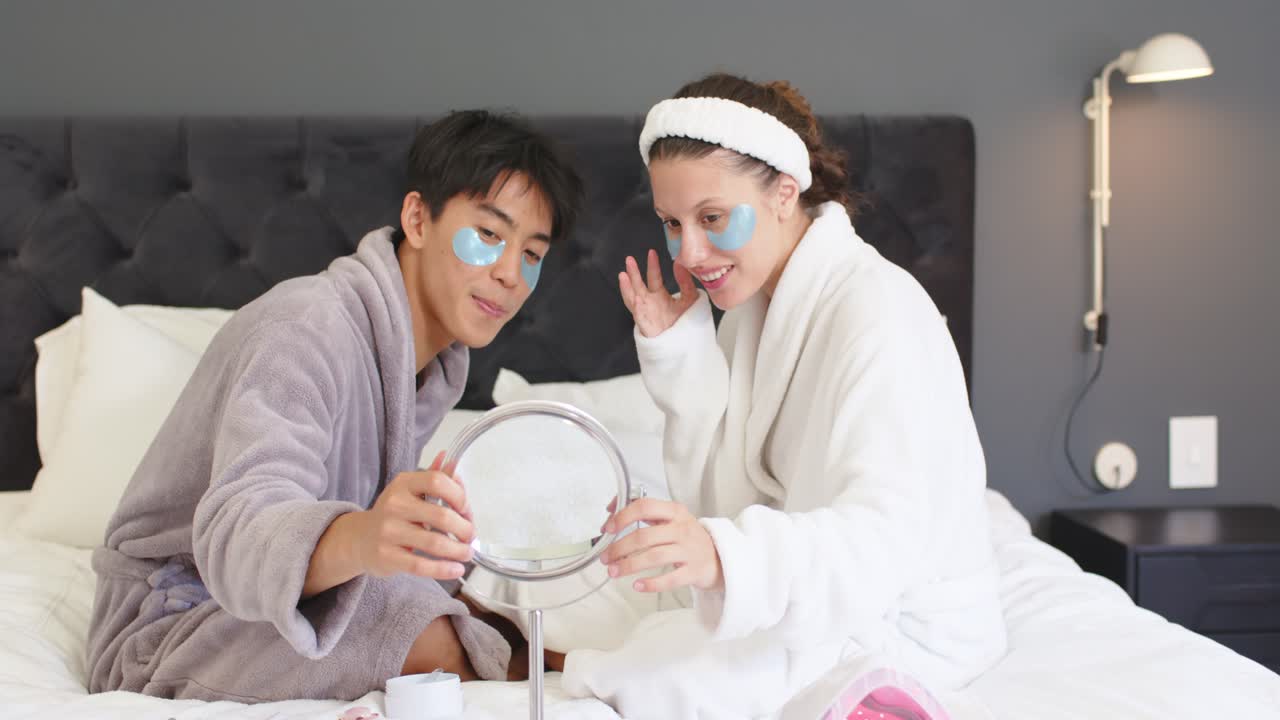 Couple in bathrobes on bed holding mirror, examining under-eye patches after placing for skin check