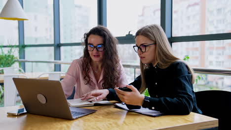 two female coworkers wearing glasses sitting at the table in office. women discussing work issues looking at the screen of laptop in front of them.