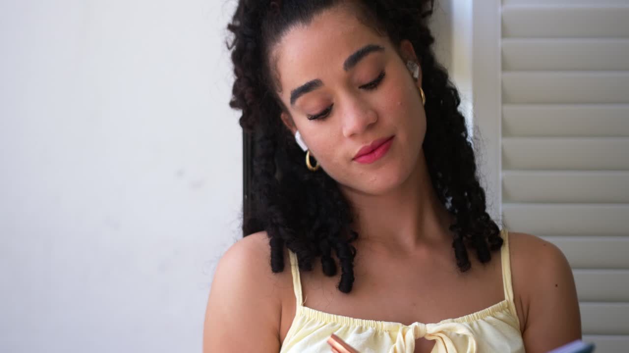 Mulher com vestido claro escrevendo com caneta ouro-rosado em um caderno azul, ouvindo com fones de ouvido na janela.