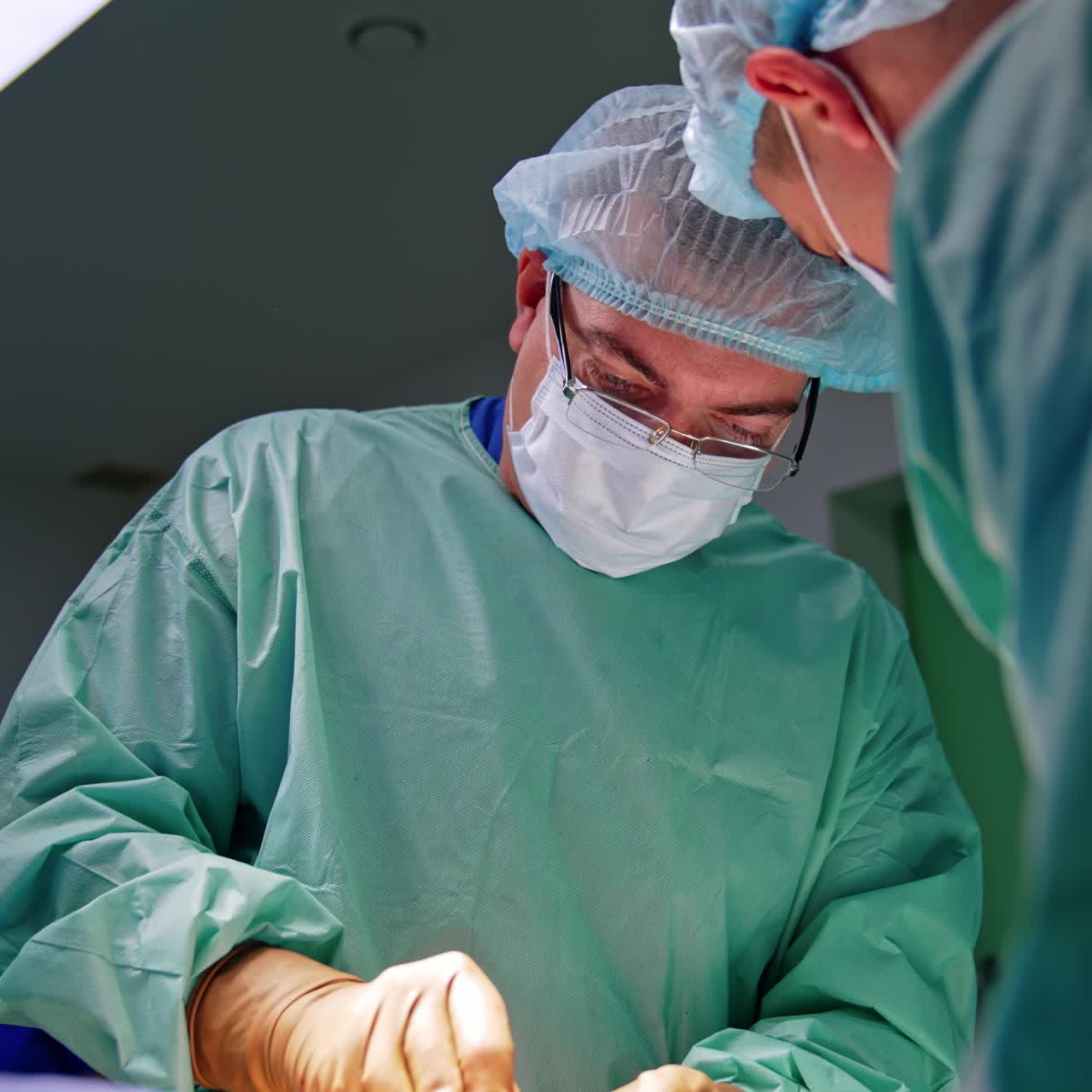 Un chirurgo adulto esperto, con cuffia e occhiali, opera. Vista dal basso sul medico impegnato nell'intervento chirurgico.