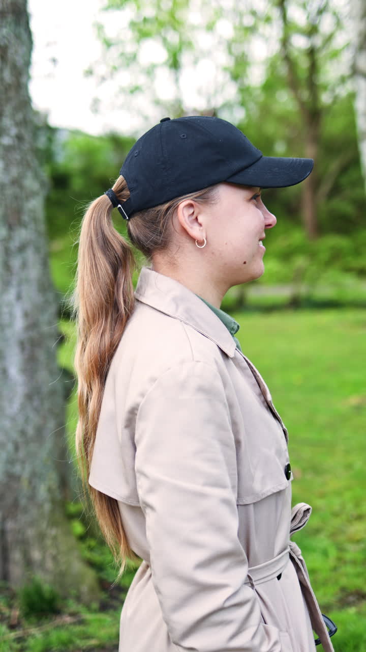 Una bella giovane donna in impermeabile passeggia lungo un sentiero in un parco, vicino agli alberi, in un ambiente tranquillo nel primo pomeriggio. Una donna graziosa a passeggio in un parco all'inizio della primavera.