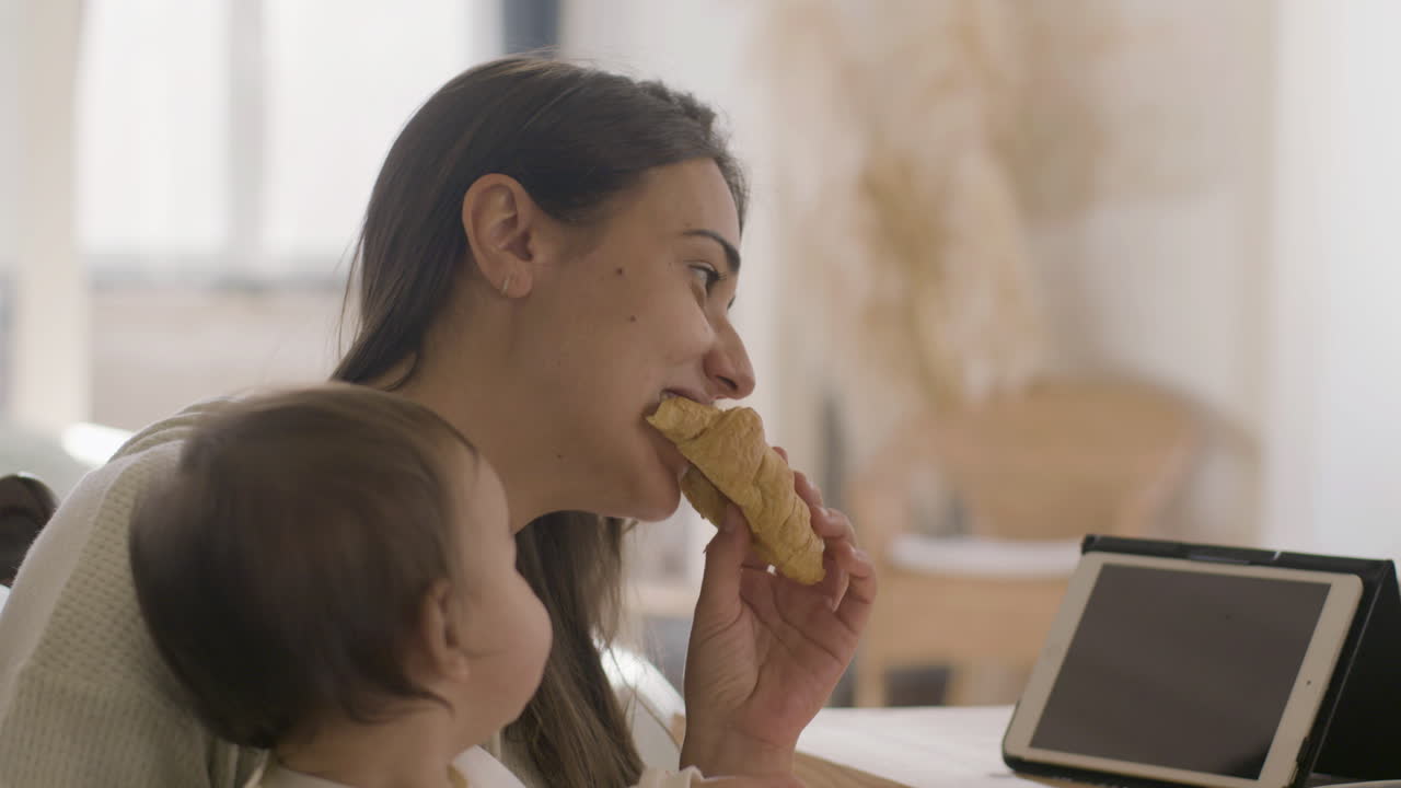 feliz mujer hermosa sentada en la mesa de la cocina y sosteniendo a su hija mientras desayunaba y comía croissant