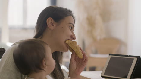 Glückliche-Schöne-Frau,-Die-Am-Küchentisch-Sitzt-Und-Ihre-Kleine-Tochter-Hält,-Während-Sie-Frühstückt-Und-Croissant-Isst