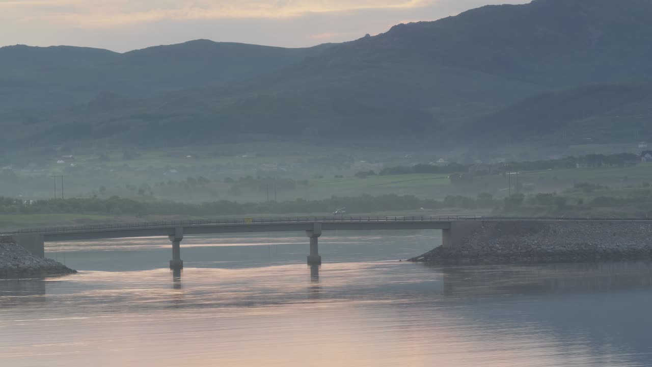 Misty summer morning. Wide angle view of mountains and lush green valley shrouded in sea mist, bridge and a passing car. Lofoten, Northern Norway