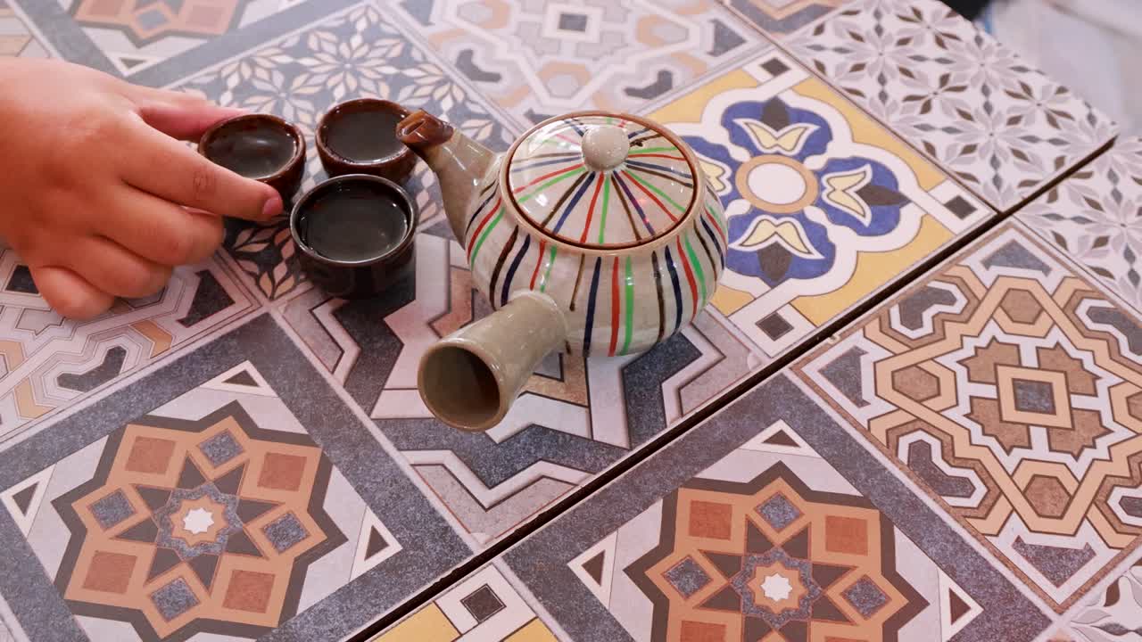 Hands pour tea from a decorative teapot into cups on a patterned tablecloth, highlighting a traditional Thai tea ceremony