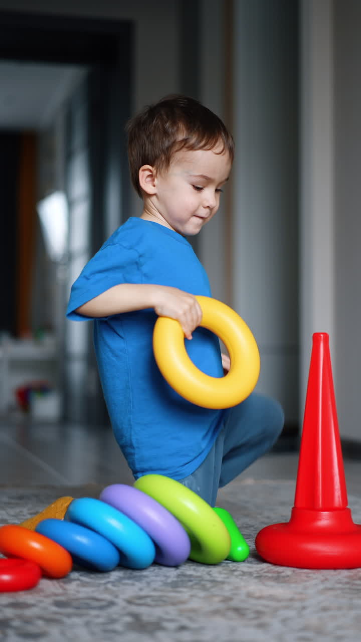 Bebé caucásico alegre sentado en el suelo sosteniendo la base de una pirámide. Niño adorable construye una pirámide en casa. Video vertical