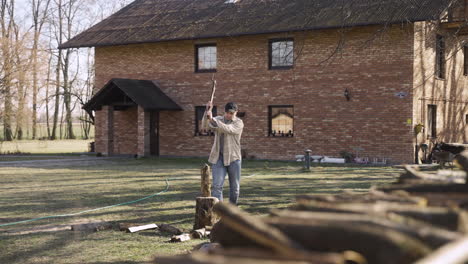 distant view of caucasian man chopping firewood with an ax outside a country house