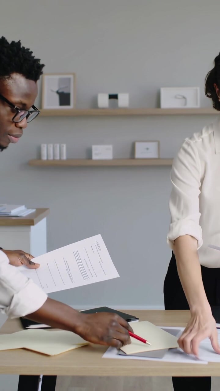 Vertical video: Leaning man in white shirt arranging files on desk, offering red pen to sign papers