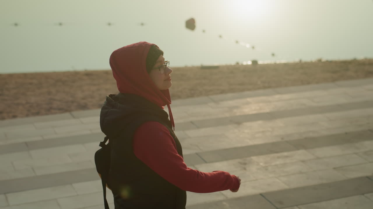 Wanita Arab yang penasaran berjalan di tepi pantai, profil tersinari matahari dalam jaket berkerudung dan ransel, langit penuh burung dan angin sepoi-sepoi pantai, berbalik cepat dan tatapan tersenyum, energi penjelajah kota yang lincah, gradasi hangat sinematik