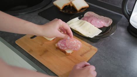hand assembling sandwiches using slices of white bread, cheese, and deli meats on wooden cutting board, highlighting preparation of simple homemade lunch in clean modern kitchen