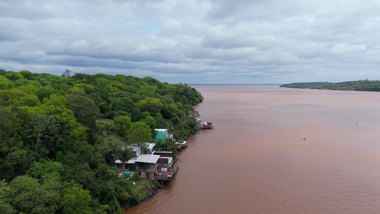 Drone glide over the Paraná River near Puerto Lagier, Misiones, Argentina, shows calm, sediment-rich waters bordered by lush forest and riverside houses nestled among dense subtropical vegetation