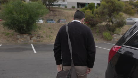 un oficinista masculino recupera su bolso de la parte trasera de su auto y lo coloca sobre su hombro mientras cruza un estacionamiento de autos hacia la oficina de su lugar de trabajo