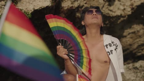 Man holding a rainbow fan and pride flag on the beach