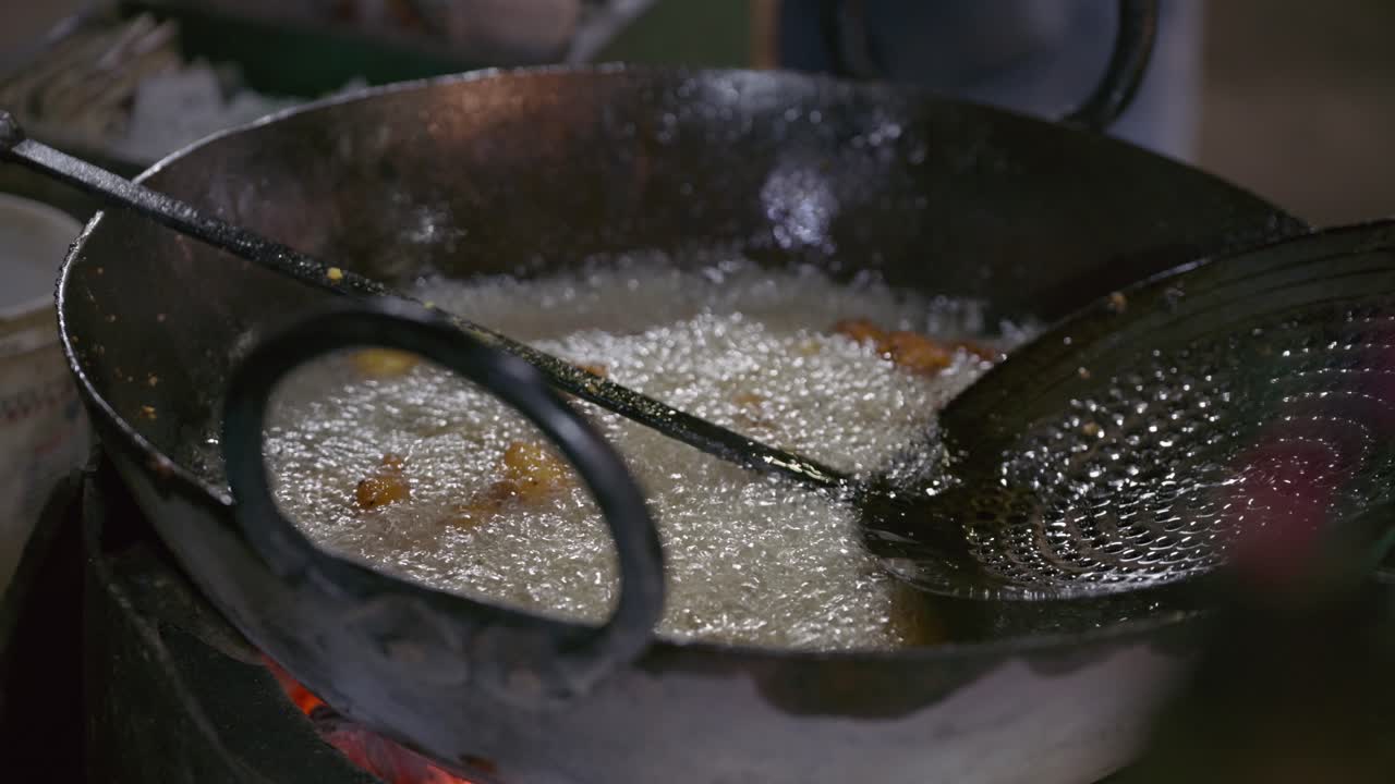Detailed close view of bhajiya Indian fritters frying in bubbling hot oil inside large wok, metal strainer in use, active street food cooking process during night
