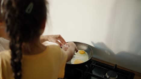 Rear-view-of-a-little-brunette-girl-in-a-yellow-dress-together-with-her-mother-preparing-fried-eggs-in-a-frying-pan-on-the-stove-in-a-modern-kitchen.-Girl-with-her-mom-learning-to-cook-breakfast,-family-morning-in-modern-apartment