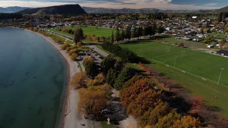 Drones-Sobrevuelan-La-Orilla-Del-Lago-Wanaka-Y-El-área-De-Recreación-Hasta-La-Ciudad,-Durante-La-Temporada-De-Otoño