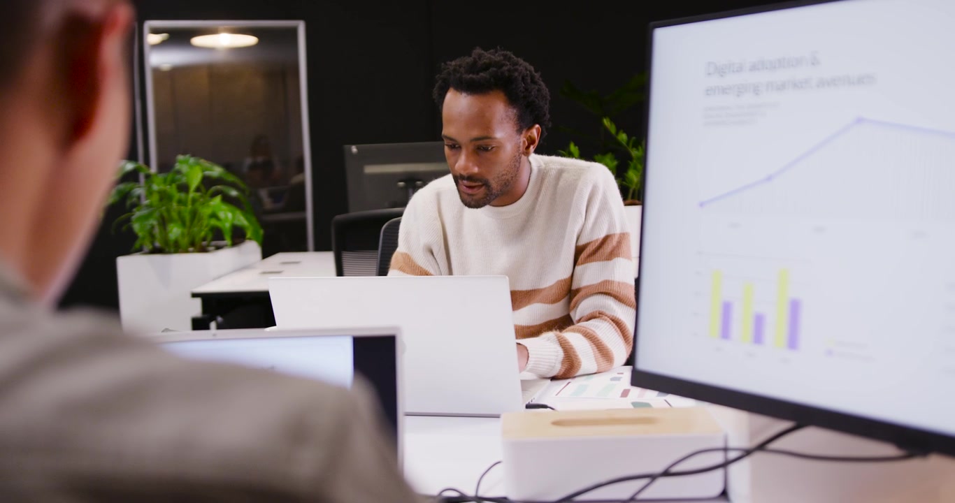 Diverse male coworkers, man of African descent checking charts, typing on laptop for work at desk