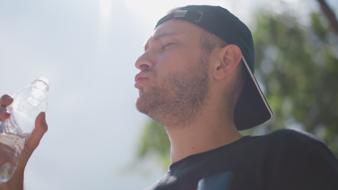 Hombre bebe al aire libre, hombre en un parque con gorra hacia atrás, escena soleada de un parque con un hombre bebiendo agua tranquilamente, hombre con gorra bebe agua tranquilamente en medio de un exuberante follaje bajo el sol del mediodía