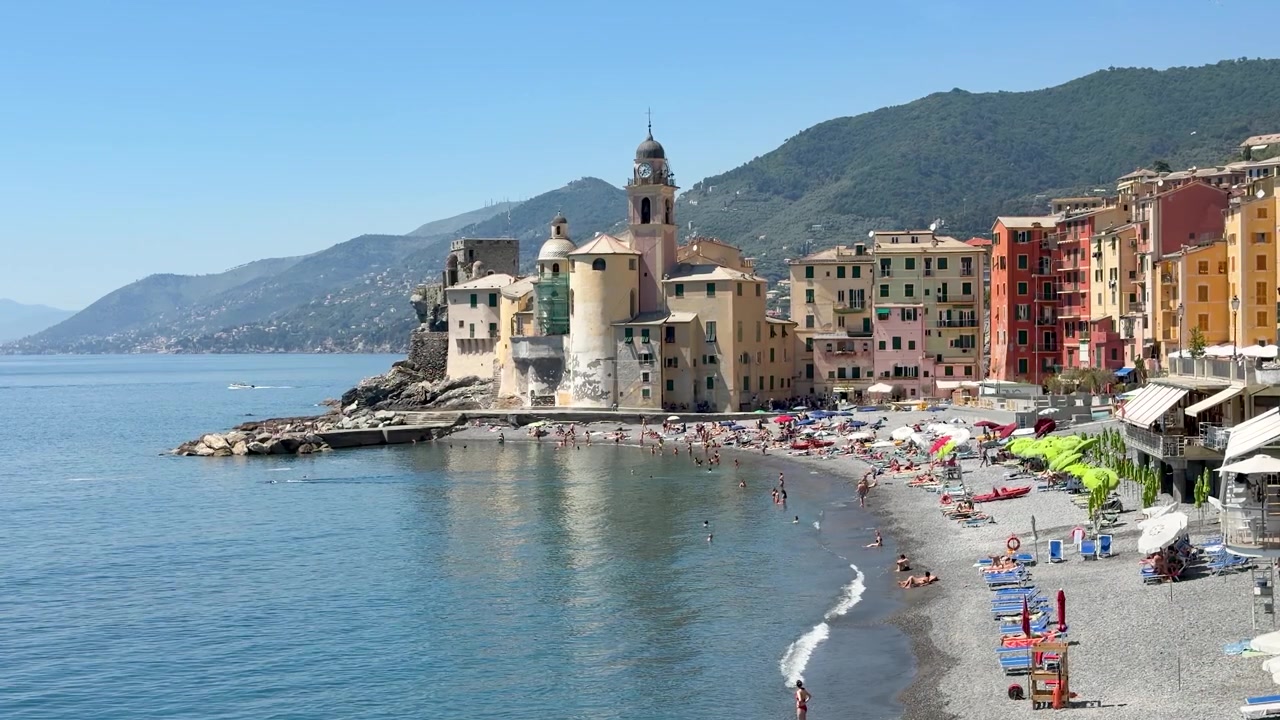 Inquadratura ampia statica di case colorate, campanile e spiaggia di ciottoli sulla costa ligure.