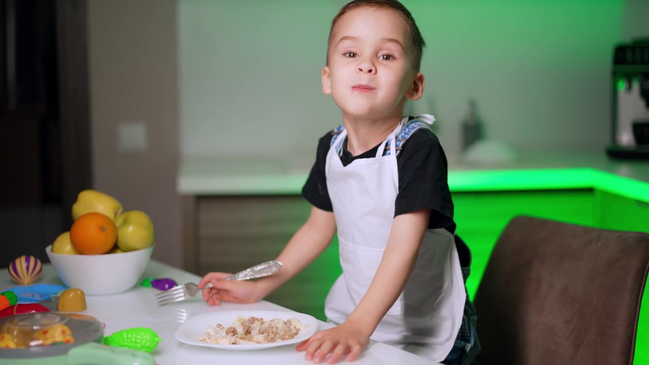 Giovane ragazzo con grembiule che tiene una forchetta e guarda la fotocamera. Un bambino è seduto a un tavolo bianco con un piatto di cibo e una ciotola di frutta sotto un'illuminazione verde in una cucina moderna.