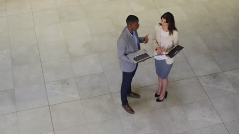 Diverse-businessman-and-businesswoman-talking-and-using-laptop-in-lobby-of-modern-office