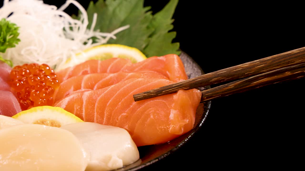 Chopsticks delicately lift a slice of salmon sashimi from a vibrant sushi platter with roe, scallop, and garnish, under bright studio lighting
