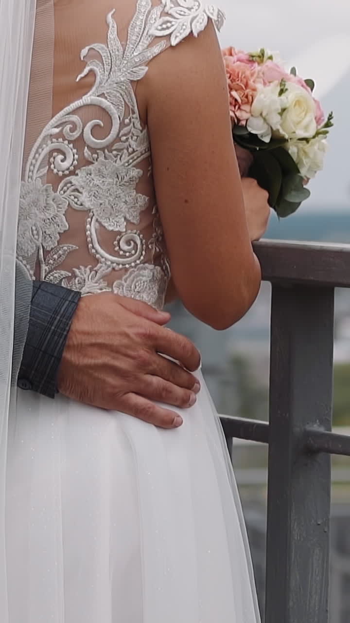 Man groom embraces beloved woman waist wearing gorgeous wedding dress standing on bridge closeup slow motion. Newlywed young bride and groom on city street