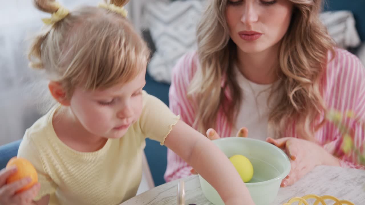 Bambina e madre preparano insieme uova di Pasqua colorate. La bambina si concentra intensamente sull'immersione di un uovo. La madre sorride calorosamente, guidando la sua bambina. La scena cattura un momento gioioso e tenero di legame familiare.